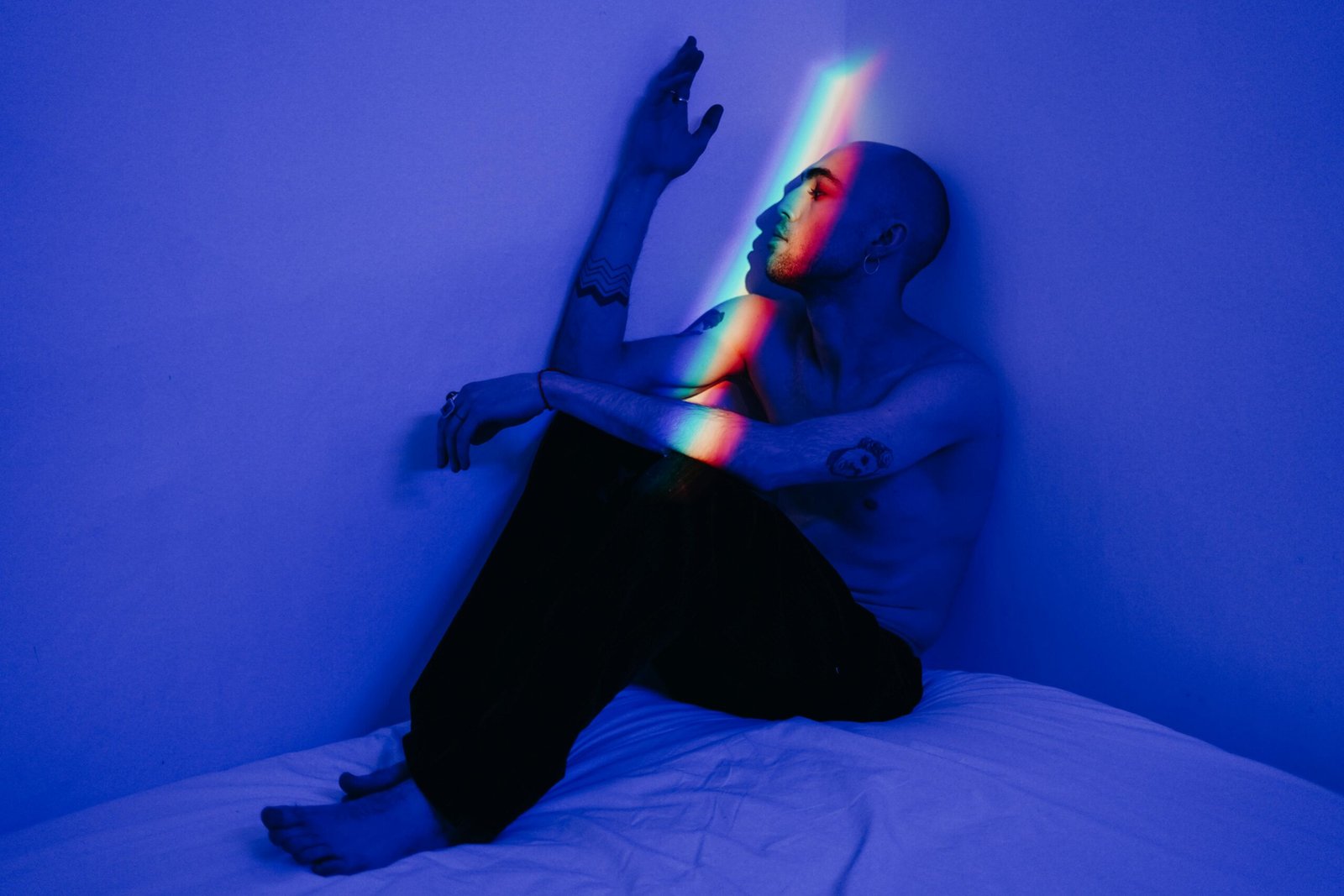 A shirtless man sitting in a blue-lit room, accented by rainbow light reflections creating a contemplative mood.